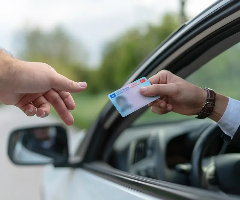 Das Bild zeigt eine Nahaufnahme von zwei Händen, die sich durch das offene Fenster eines Autos begegnen. Eine Hand von außerhalb des Autos greift nach einem Ausweis oder Führerschein, der von der Hand einer Person im Auto übergeben wird. Die Übergabe findet in einer grünen, unscharf abgebildeten Umgebung statt. Die Szene vermittelt den Moment der Kontrolle oder Übergabe eines wichtigen Dokuments, möglicherweise im Zusammenhang mit einer Fahrprüfung oder Verkehrskontrolle.