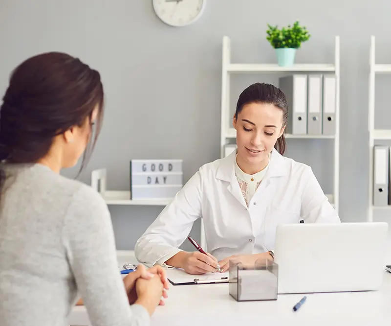 Eine Frau in professioneller Kleidung sitzt an einem Schreibtisch, schreibt in ein Notizbuch und lächelt freundlich. Auf der anderen Seite des Schreibtischs sitzt eine weitere Person, deren Gesicht nicht sichtbar ist. Der Raum ist hell und modern eingerichtet, im Hintergrund sind Regale mit Aktenordnern und eine kleine Pflanze zu sehen.