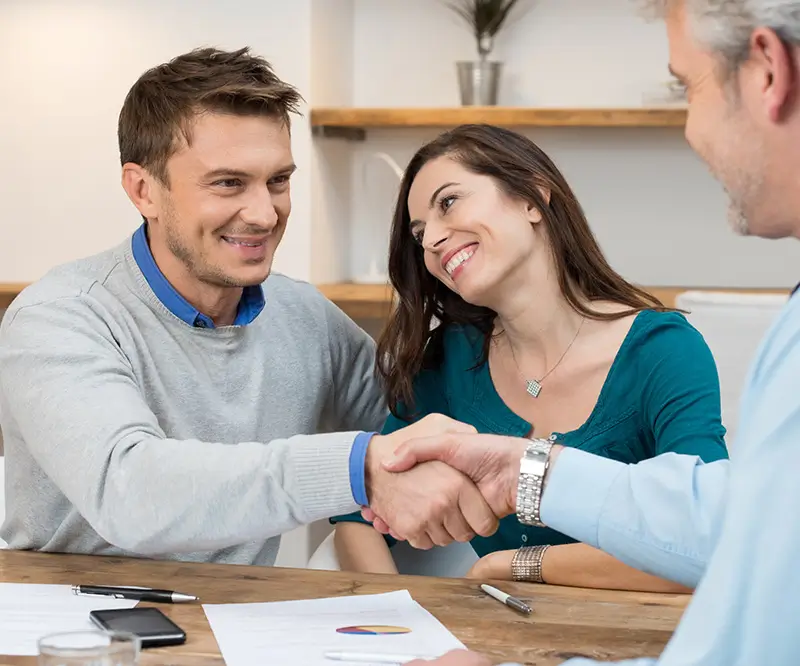 Ein lächelndes Paar sitzt an einem Tisch und gibt einem Mann, der ebenfalls lächelt, die Hand. Auf dem Tisch liegen Dokumente und Stifte. Die Szene vermittelt ein Gefühl von Einigung und Zufriedenheit, symbolisierend den positiven Abschluss einer Beratung oder eines Geschäftsgesprächs.