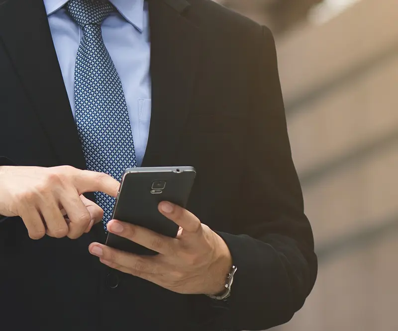 Das Bild zeigt den Oberkörper eines Mannes in einem Anzug, der ein Smartphone in den Händen hält. Der Mann trägt ein hellblaues Hemd und eine gemusterte Krawatte. Mit einem Finger tippt er auf den Bildschirm des Smartphones, als ob er eine Nachricht schreibt oder eine Nummer wählt. Der Hintergrund ist unscharf, und die Szene scheint draußen in einem städtischen Umfeld aufgenommen worden zu sein. Das Bild vermittelt Professionalität und Kommunikation.