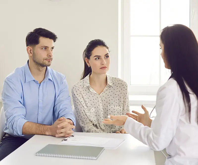 Das Bild zeigt ein ernsthaftes Gespräch zwischen drei Personen in einem hellen, neutral eingerichteten Raum. Auf der linken Seite sitzen ein Mann und eine Frau nebeneinander am Tisch. Beide schauen konzentriert auf die dritte Person, eine Frau im weißen Kittel, die ihnen gegenüber sitzt und mit den Händen gestikuliert. Vor dem Paar liegt ein Notizbuch auf dem Tisch. Die Szene deutet auf eine Beratung oder ein wichtiges Gespräch hin, möglicherweise im Zusammenhang mit einer MPU-Beratung oder psychologischen Unterstützung.