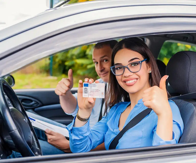 Das Bild zeigt eine junge Frau mit dunklem Haar und Brille, die auf dem Fahrersitz eines Autos sitzt. Sie lächelt fröhlich und hält ihren Führerschein in der rechten Hand hoch, während sie mit der linken Hand einen Daumen nach oben zeigt. Neben ihr, auf dem Beifahrersitz, sitzt ein Mann, der ebenfalls lächelt und einen Daumen nach oben zeigt, möglicherweise ein Fahrprüfer oder Berater. Beide wirken zufrieden und stolz, als ob die Frau gerade ihre Fahrprüfung bestanden oder ihren Führerschein zurückerhalten hat. Die Szene findet in einem Auto statt, das in einer grünen Umgebung geparkt ist.