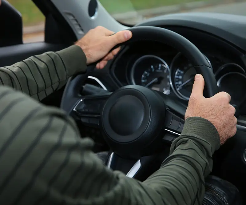 Eine Person, die ein grünes, gestreiftes langärmeliges Hemd trägt, hält das Lenkrad eines Autos und konzentriert sich auf das Fahren. Das Armaturenbrett und die Bedienelemente sind sichtbar, was darauf hindeutet, dass die Person aktiv am Fahrvorgang beteiligt ist.