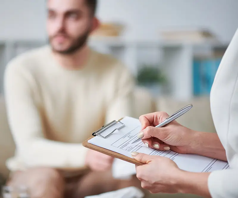 Eine Person schreibt mit einem Stift auf einem Klemmbrett, während im Hintergrund ein unscharf abgebildeter Mann sitzt. Die Szene deutet auf ein Beratungsgespräch oder eine therapeutische Sitzung hin