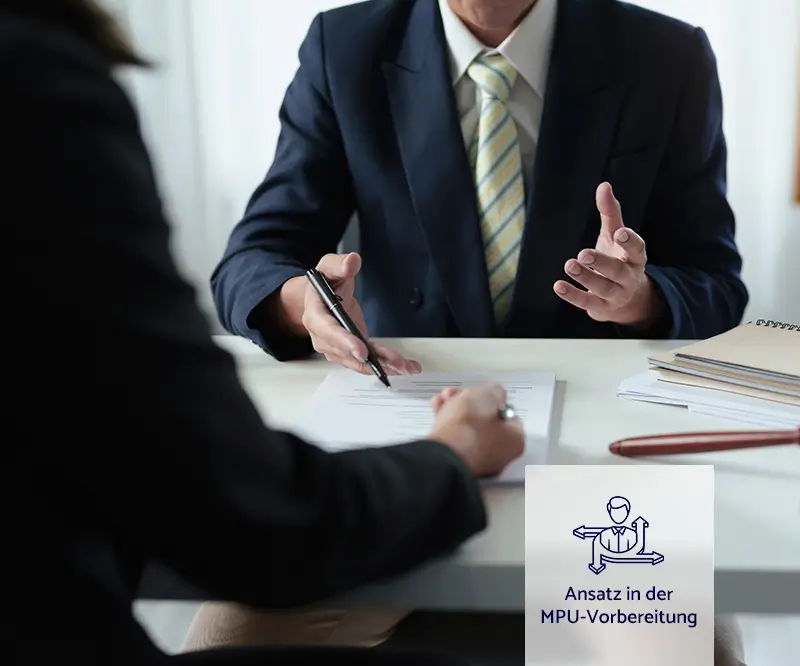 Das Bild zeigt ein Gespräch zwischen zwei Personen, die an einem Tisch sitzen. Der Fokus liegt auf den Händen und Oberkörpern der Personen. Eine Person, die einen dunklen Anzug und eine gestreifte Krawatte trägt, hält einen Stift in der Hand und scheint etwas zu erklären oder zu unterzeichnen. Die andere Person sitzt gegenüber und hört aufmerksam zu, während sie ebenfalls einen Stift hält. Auf dem Tisch liegen Dokumente und Notizbücher. In der unteren rechten Ecke des Bildes befindet sich ein Icon mit einer stilisierten Figur und der Text „Ansatz in der MPU-Vorbereitung“.