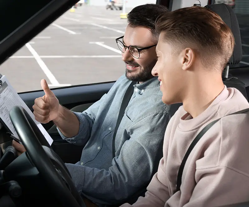 Das Bild zeigt eine Szene in einem Auto, in der ein Fahrlehrer und ein Fahrschüler auf den Prüfungsbogen schauen. Der Fahrlehrer, ein Mann mit Brille, hebt den Daumen, um dem Schüler zu signalisieren, dass alles gut läuft. Der Fahrschüler, ein junger Mann in einem hellen Pullover, lächelt und wirkt erleichtert. Die beiden sitzen auf einem Parkplatz, was durch die leeren Parkplätze im Hintergrund erkennbar ist. Die Szene vermittelt ein Gefühl von Unterstützung und Erfolg während einer Fahrprüfung.