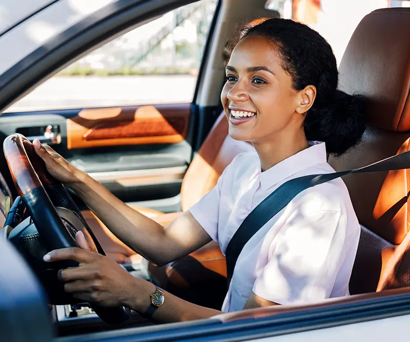 Eine lächelnde Frau mit dunklem Haar, das zu einem lockeren Dutt gebunden ist, sitzt in einem Auto und hält das Lenkrad mit beiden Händen. Sie trägt einen Sicherheitsgurt und scheint während der Fahrt glücklich und konzentriert zu sein. Der Innenraum des Autos wirkt hochwertig, mit braunen Lederpolstern.