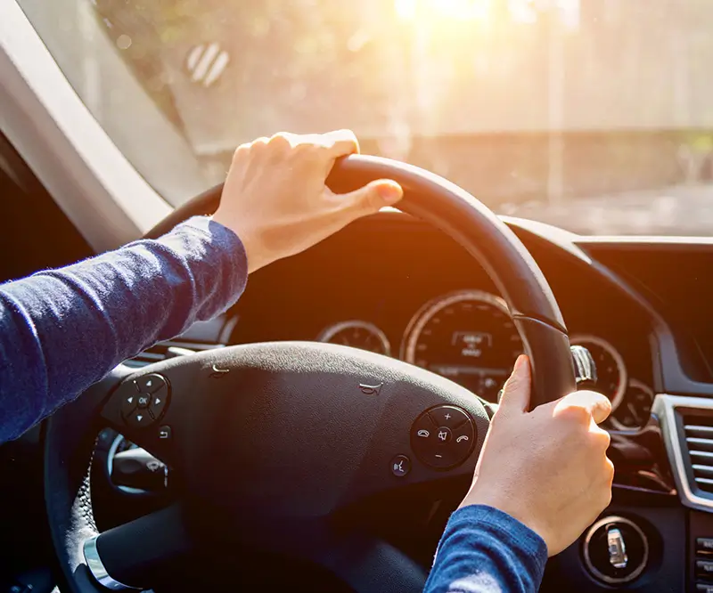 Das Bild zeigt eine Nahaufnahme von zwei Händen, die das Lenkrad eines Autos halten. Der Fahrer trägt ein langärmeliges blaues Oberteil, und durch die Windschutzscheibe ist ein sonniger, leicht unscharfer Hintergrund zu sehen. Das Bild vermittelt ein Gefühl von Kontrolle und Fokussierung während der Fahrt.