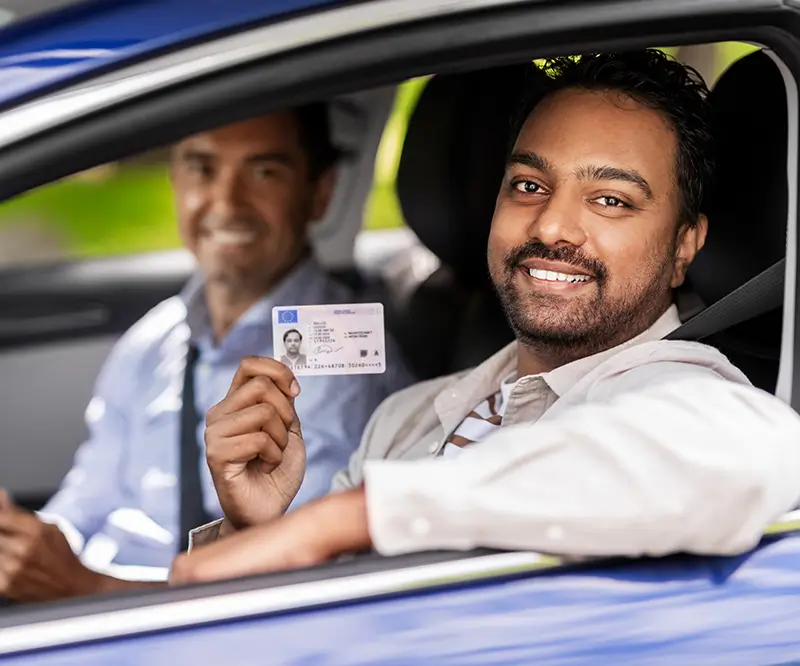 Das Bild zeigt einen Mann mit dunklen Haaren und Bart, der lächelnd im Fahrersitz eines Autos sitzt und seinen Führerschein in die Kamera hält. Neben ihm, auf dem Beifahrersitz, sitzt ein weiterer Mann, der ebenfalls lächelt. Beide tragen Sicherheitsgurte, und die Stimmung wirkt freudig und erleichtert, als ob der Fahrer gerade seinen Führerschein zurückerhalten hat.