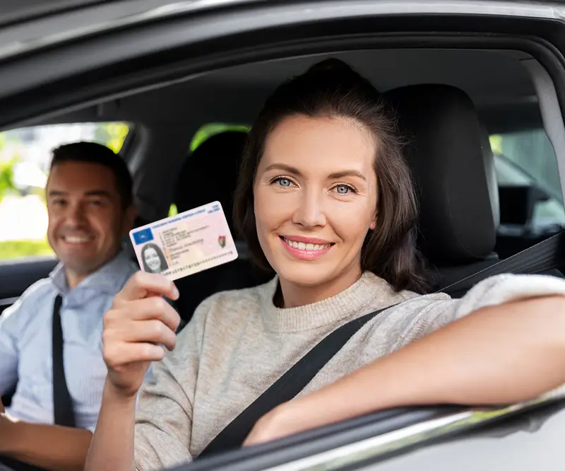 Eine Frau mit braunen Haaren, die auf dem Fahrersitz eines Autos sitzt und lächelnd ihren Führerschein hochhält. Sie trägt einen hellen Pullover. Im Hintergrund sitzt ein Mann auf dem Beifahrersitz, der ebenfalls lächelt. Das Bild vermittelt ein Gefühl von Erfolg und Zufriedenheit, möglicherweise im Zusammenhang mit dem Bestehen einer Fahrprüfung oder dem Wiedererwerb des Führerscheins.